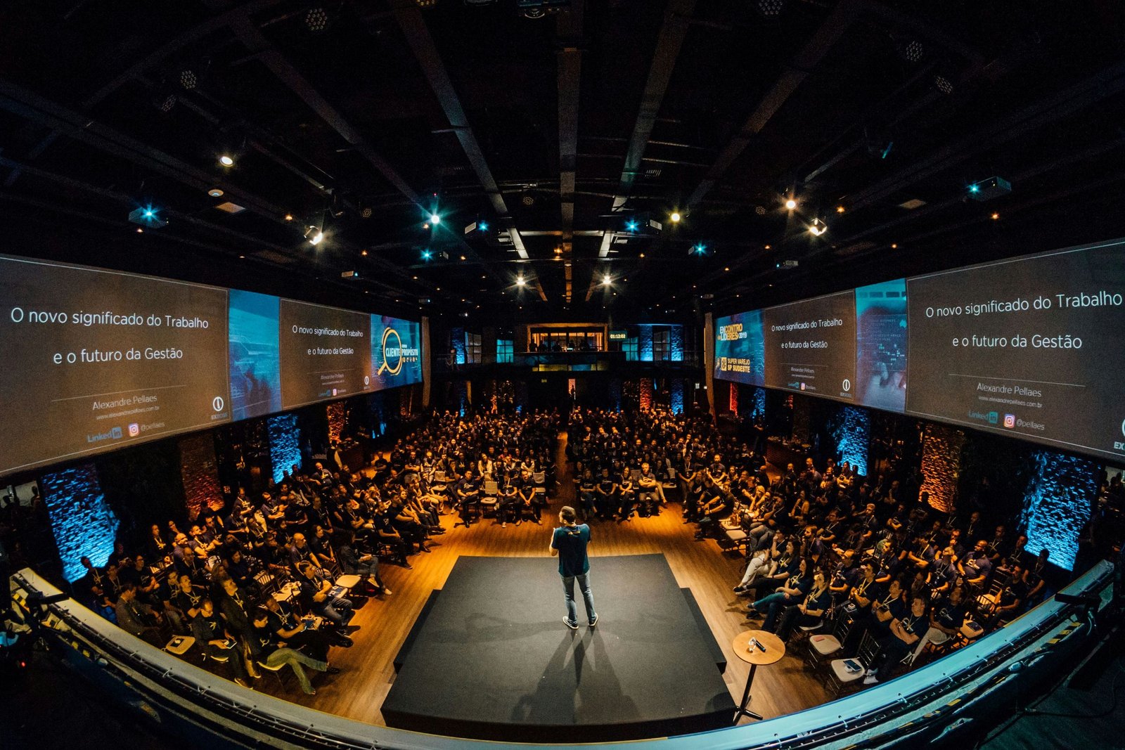 A speaker presenting on a lit stage to a full conference audience, with wide screens on both sides of the hall