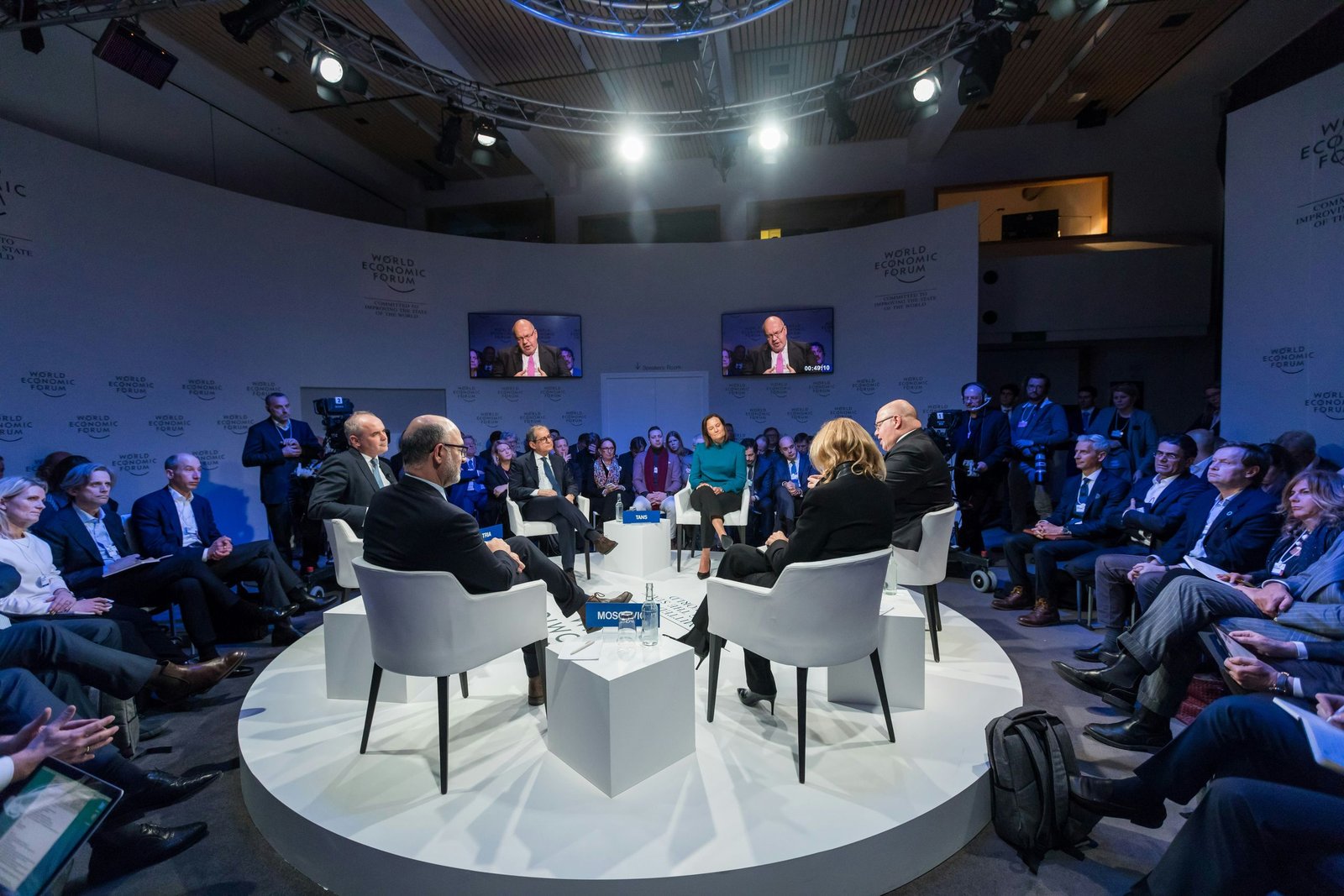 Panelists seated in white chairs on a round stage with audience around them